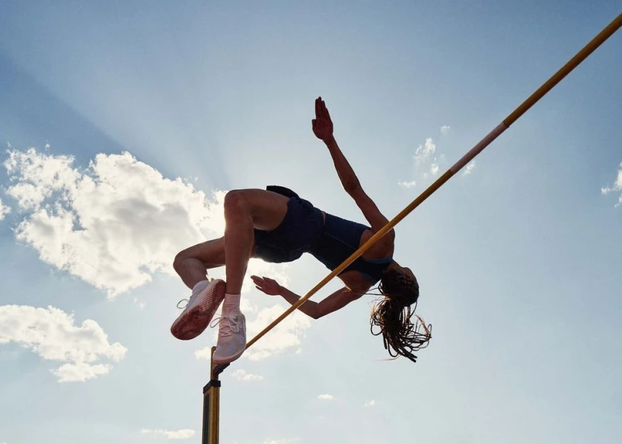 High angle view of a female athlete performing a high jump over a yellow bar against a blue sky with clouds and sun rays.
