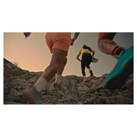 Low-angle shot of group running on rocky terrain during daytime, wearing athletic wear and colorful athletic shoes, against a sunset sky.