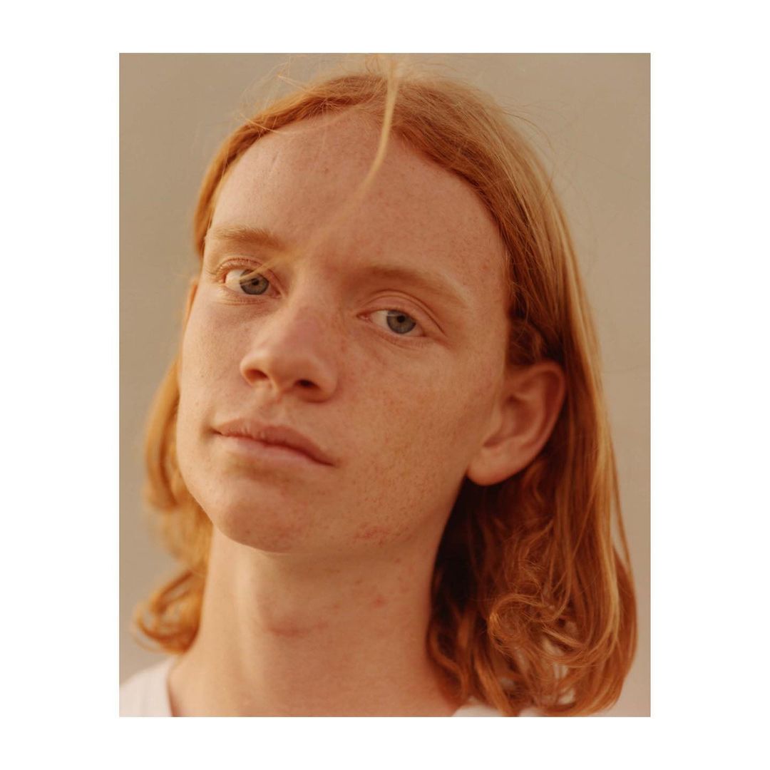 Close-up portrait of a young adult with blue eyes and ginger hair, looking directly at the camera against a neutral studio backdrop.