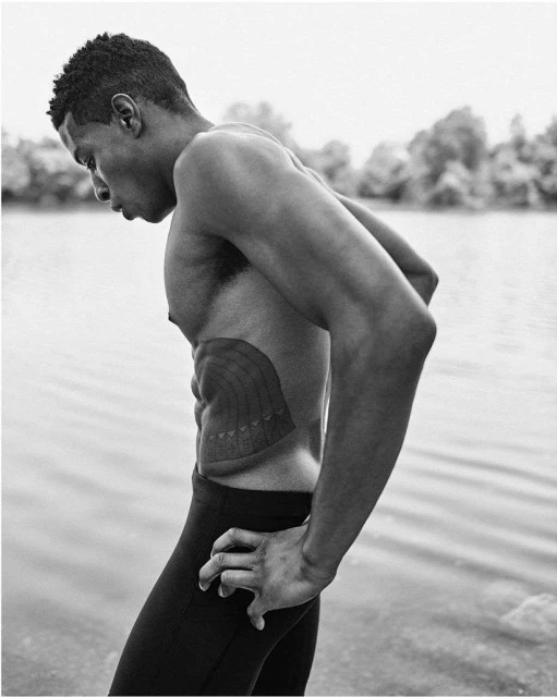 Athletic man in black leggings standing by a body of water. Trees are visible in the background, creating a serene, monochromatic scene.