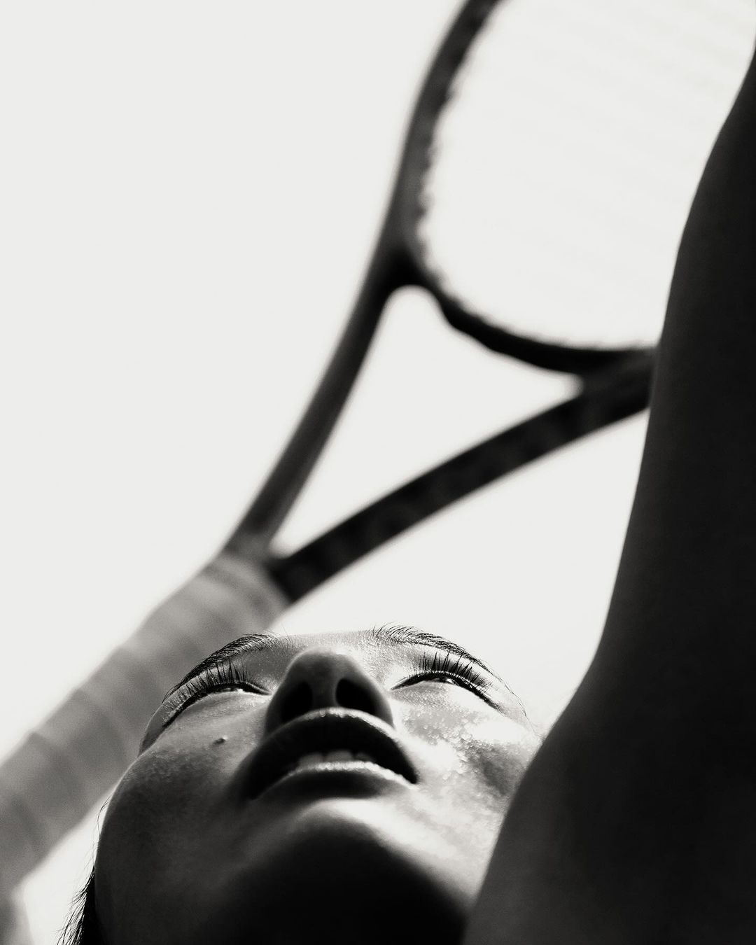 Low-angle, black-and-white portrait of a sweaty woman's face looking up, with a tennis racket out of focus in the background against a white sky.