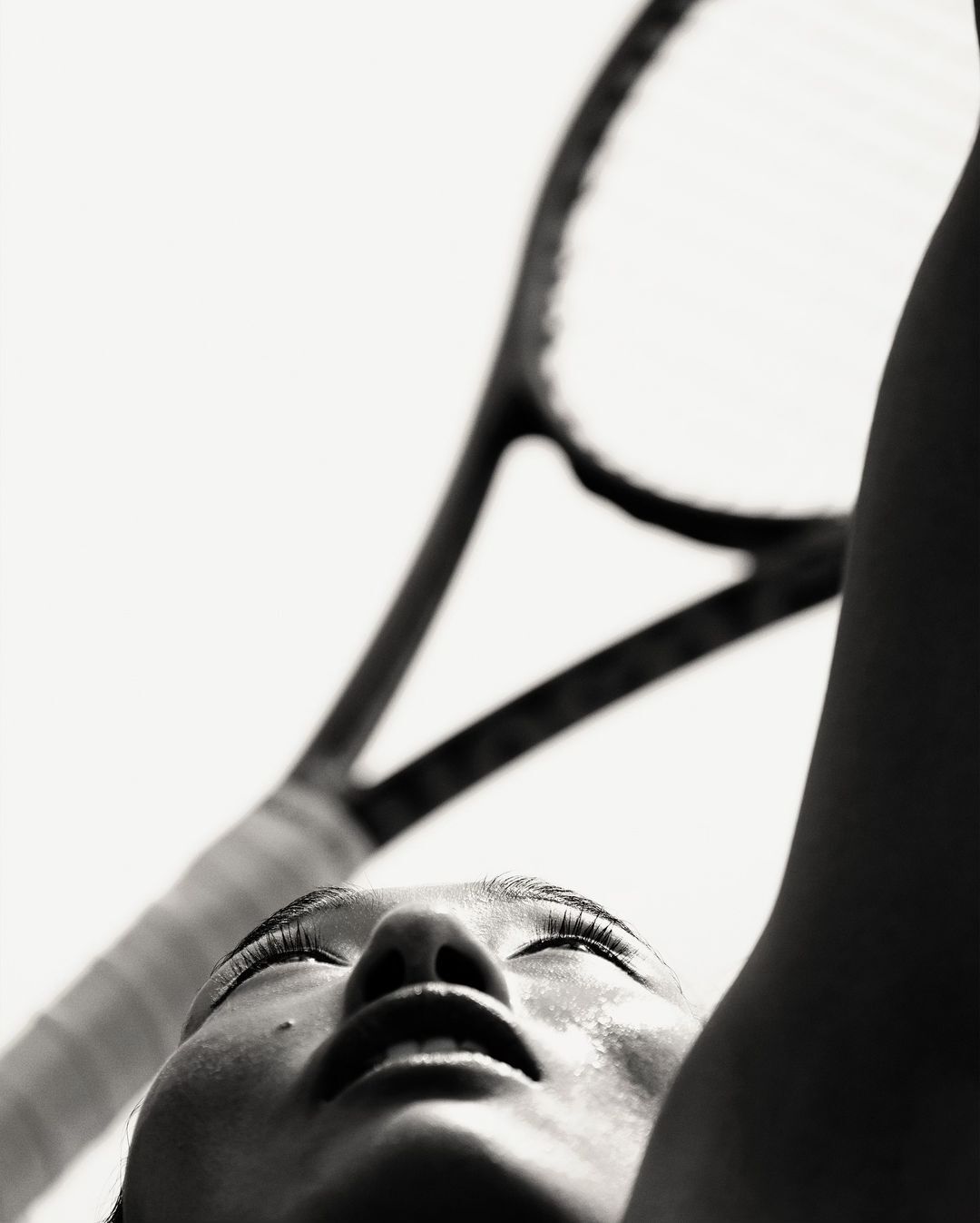 Close-up shot in black and white of an adult female looking up at a tennis racket with sweat on her face, captured with a low angle.