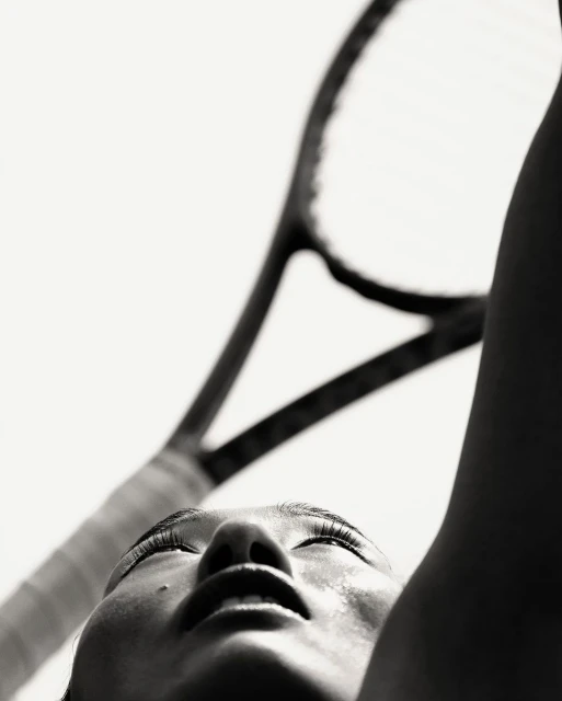 Close-up shot in black and white of an adult female looking up at a tennis racket with sweat on her face, captured with a low angle.