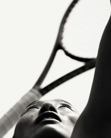 Close-up shot in black and white of an adult female looking up at a tennis racket with sweat on her face, captured with a low angle.