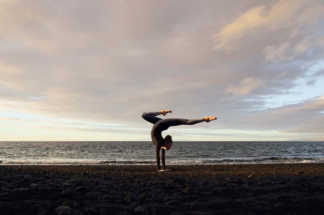 Woman in a dark bodysuit performs a handstand on a black sand beach near the ocean beneath a cloudy sky at dusk or dawn.