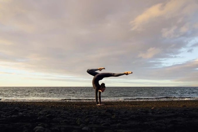 Woman in a dark bodysuit performs a handstand on a black sand beach near the ocean beneath a cloudy sky at dusk or dawn.