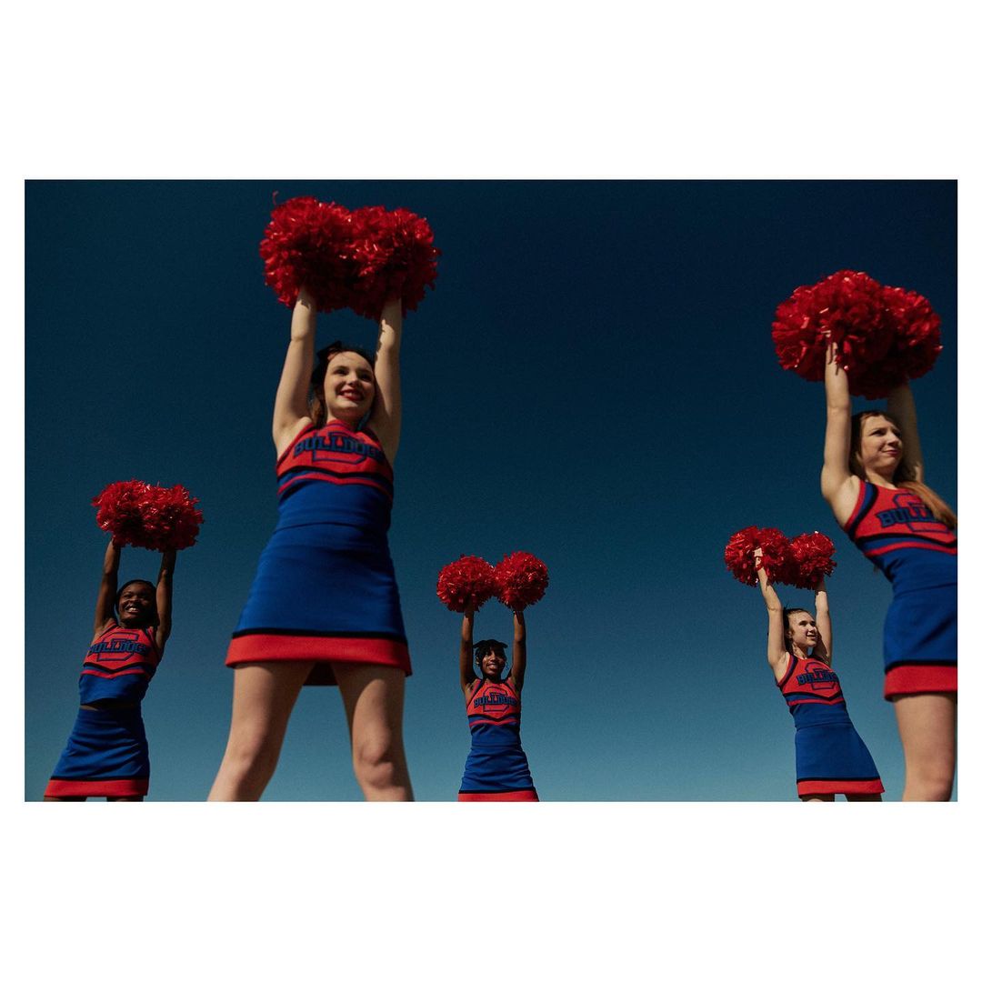 Five cheerleaders in blue and red uniforms raise red pom-poms against a clear blue sky. The cheerleaders are positioned in an outdoor setting.