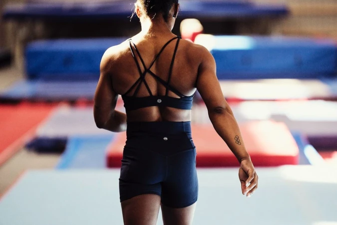 Athletic woman in workout clothes faces away from the camera, back muscles defined in a fitness setting with mats on the floor.