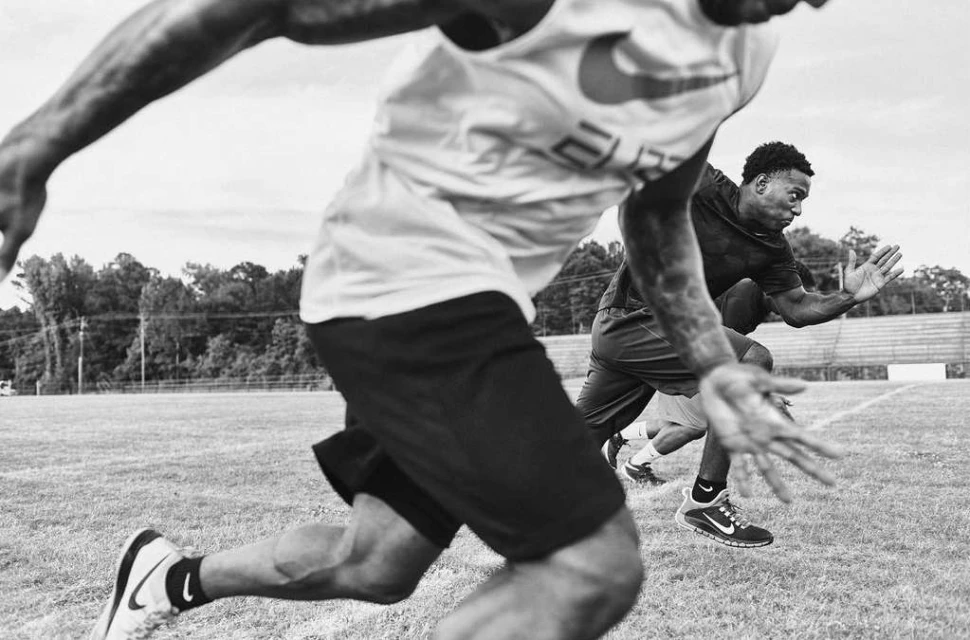 Two male athletes in athletic wear sprinting outdoors on a grassy field under an overcast sky in a black and white sports image.