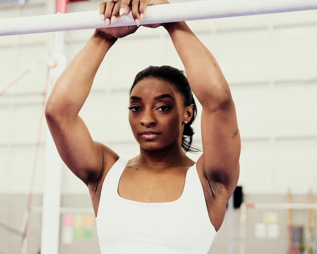 A female gymnast with tan skin in a white leotard is holding onto a white balance beam in an indoor sports training facility with natural light.