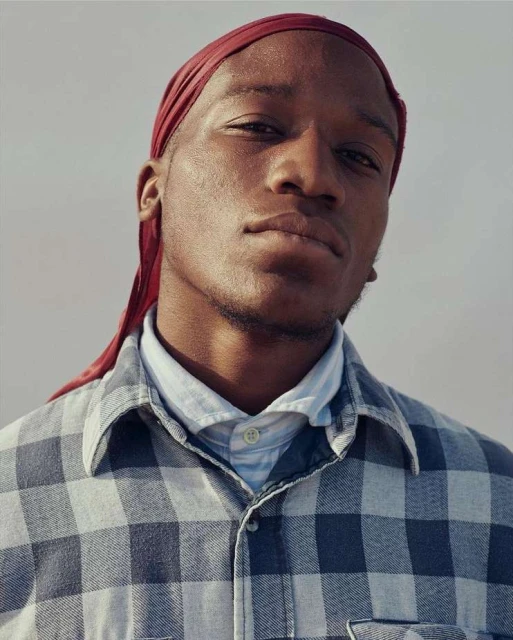Portrait of a young Black man wearing a red durag and blue plaid shirt, shot at eye-level in an indoor setting against a neutral background.