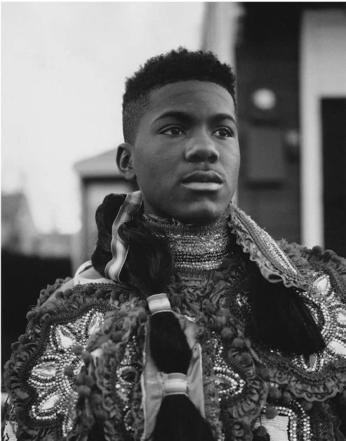 Portrait of a young Black man in ornamental clothing and braided hair, looking off-camera. Black and white photo taken outdoors.