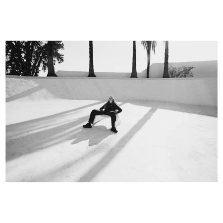 Black and white photograph of a person seated in a chair within a sunlit empty outdoor pool surrounded by palm trees.