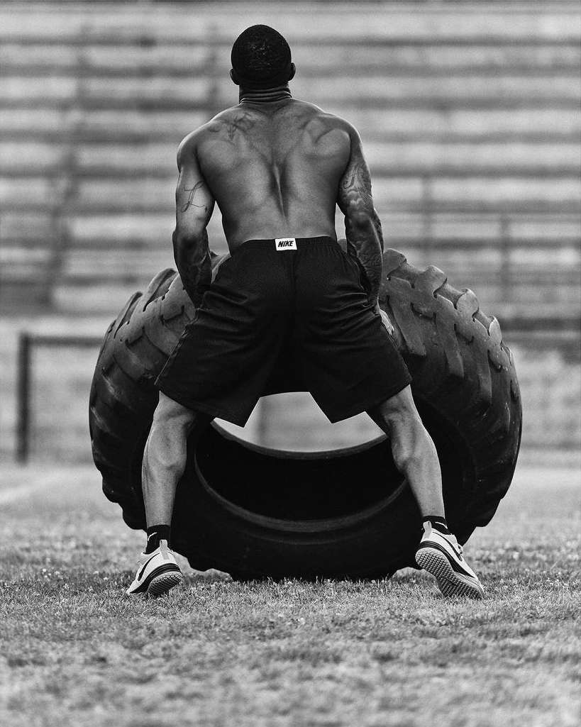 An African American adult male with tattoos pushes a large tractor tire outdoors on a grassy field, photographed in black and white.