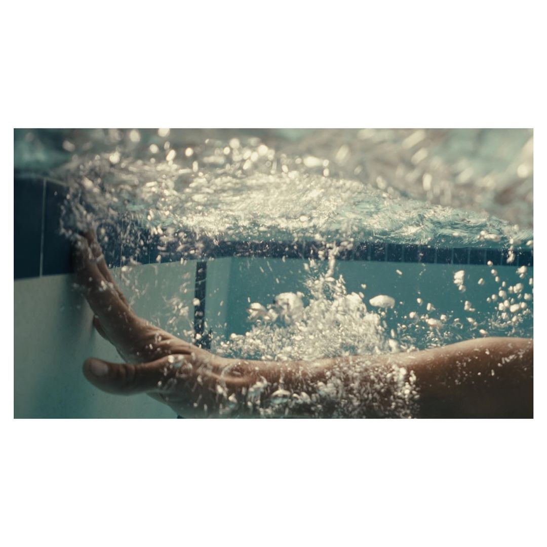 Underwater view of a person's arm and hand touching the edge of a swimming pool, with bubbles and filtered light creating a tranquil scene.