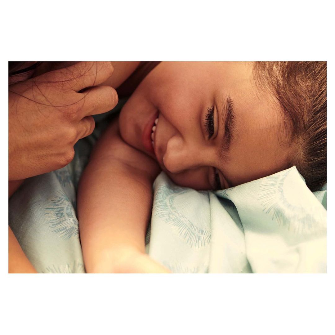 Close-up portrait of a smiling child lying on patterned light-blue bedding with an adult hand resting nearby.