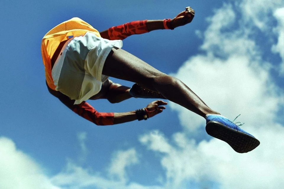An adult male with dark skin wearing an orange shirt, shorts, and blue running shoes is suspended mid-air against a sky background.