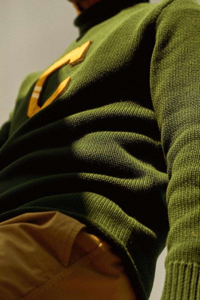 Close-up on a person wearing a green knit sweater with a large yellow letter 'C', and tan pants, showcasing knitwear texture.