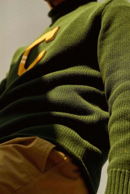 Close-up on a person wearing a green knit sweater with a large yellow letter 'C', and tan pants, showcasing knitwear texture.