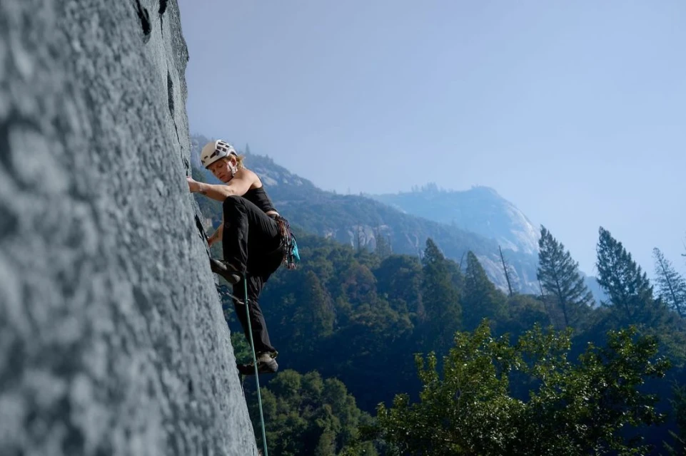 Adult female rock climber with a helmet ascends a steep, gray rock face outdoors, trees and mountain in background on a sunny day.