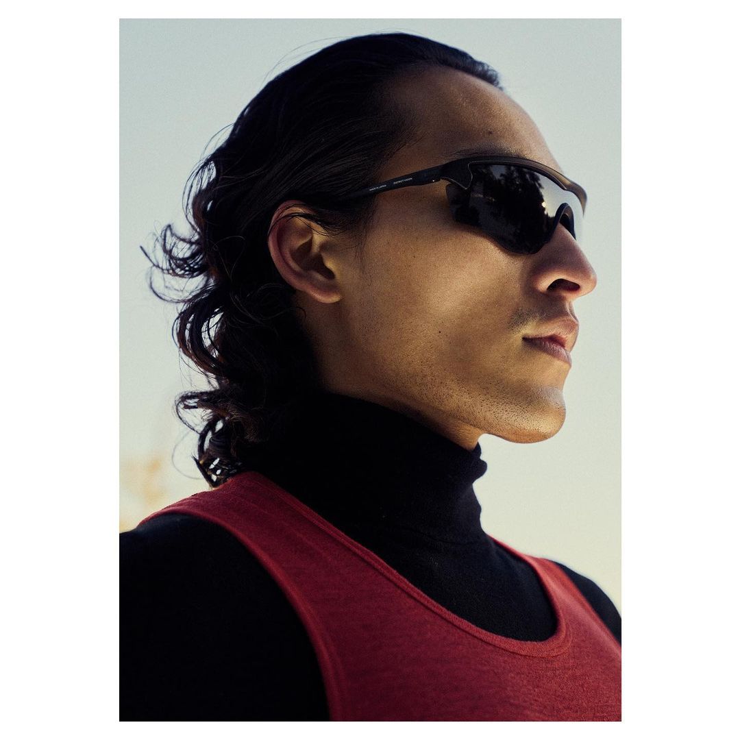 Portrait of an adult male wearing sunglasses, a black turtleneck, and a red tank top against a bright, neutral outdoor background.