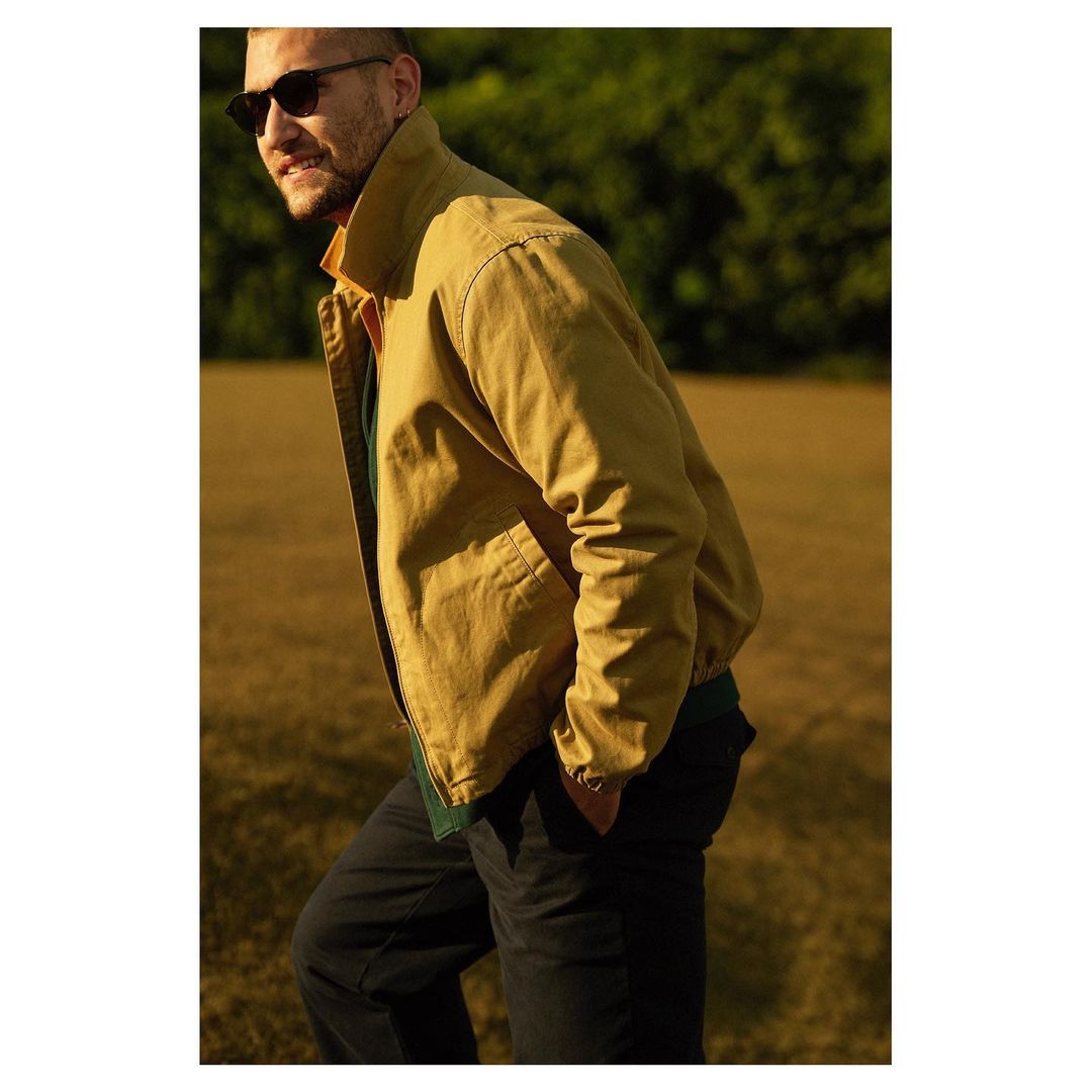 A medium shot of an adult male wearing sunglasses and a khaki jacket, standing in a grassy field with trees in the background, captured outdoors.