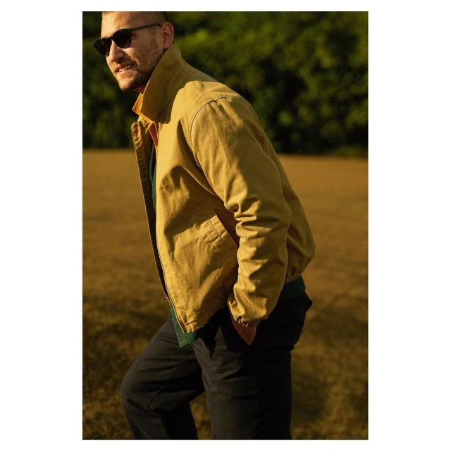 A medium shot of an adult male wearing sunglasses and a khaki jacket, standing in a grassy field with trees in the background, captured outdoors.