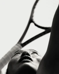 Low angle monochrome photograph of a person holding a tennis racket up against a bright sky background.