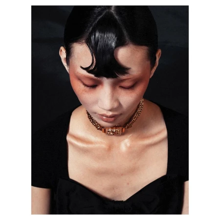 Close-up portrait of a woman wearing a heavy gold chain choker necklace and a black top against a dark background.