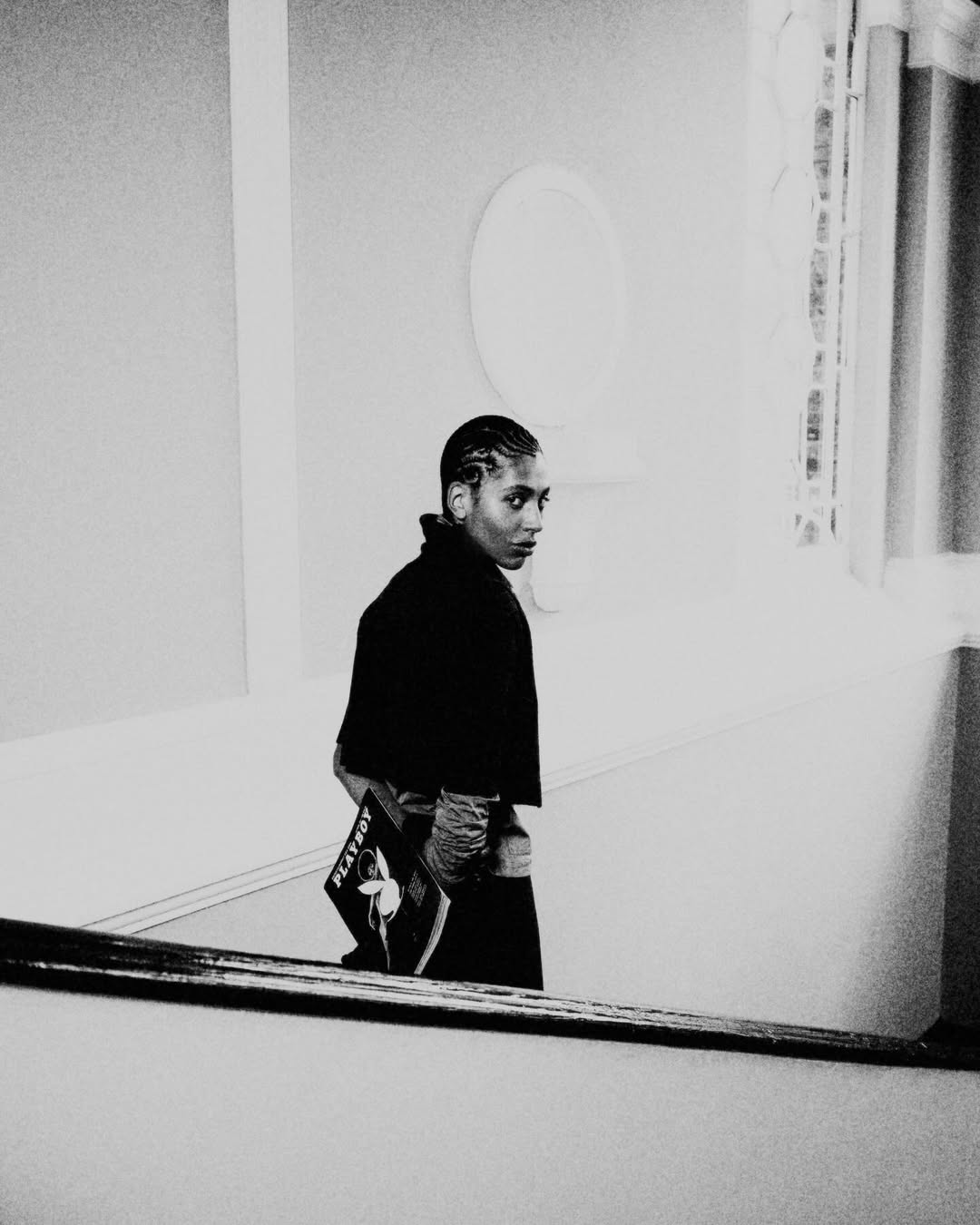 Black and white photo of a woman with short hair, looking over her shoulder while holding a magazine indoors near a window.