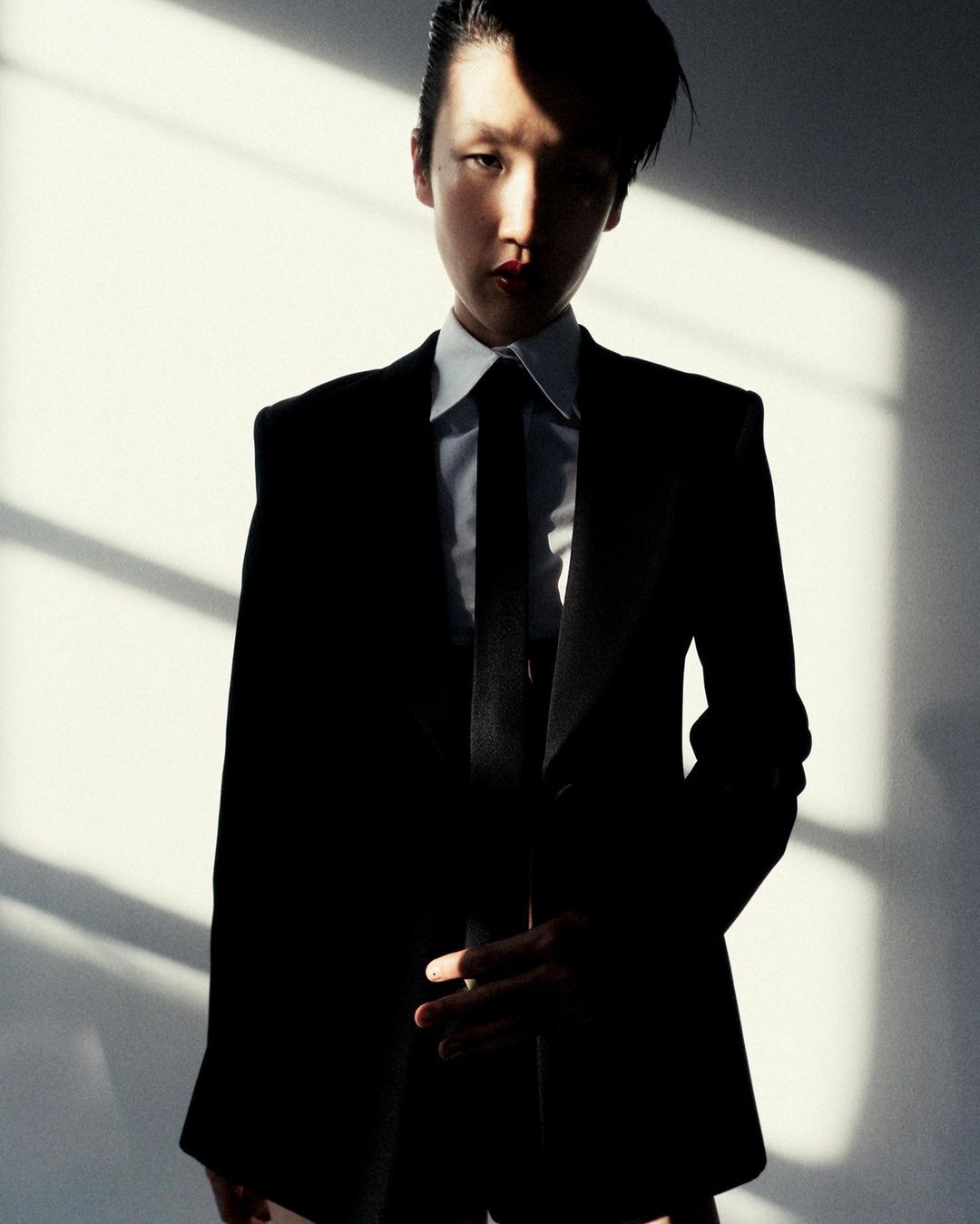 An Asian woman in a black suit and white dress shirt stands in front of a white wall with light and shadow creating diagonal lines.