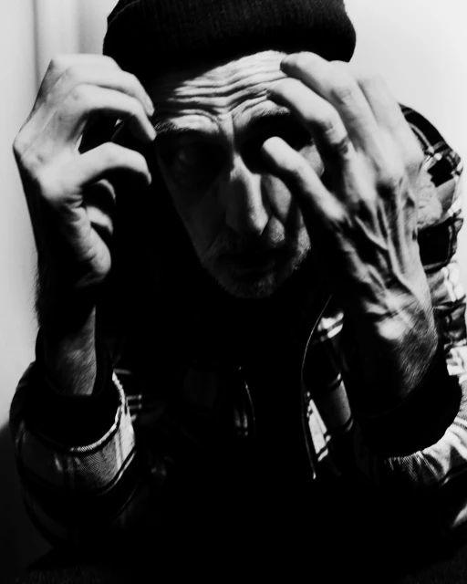 Monochrome portrait of an adult male wearing a beanie and plaid shirt, holding his hands near his face with a concerned expression.