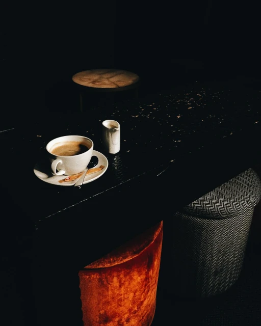 Close-up shot of a cup of coffee with a creamer on a black table, dark background with two stools adding depth to the scene.