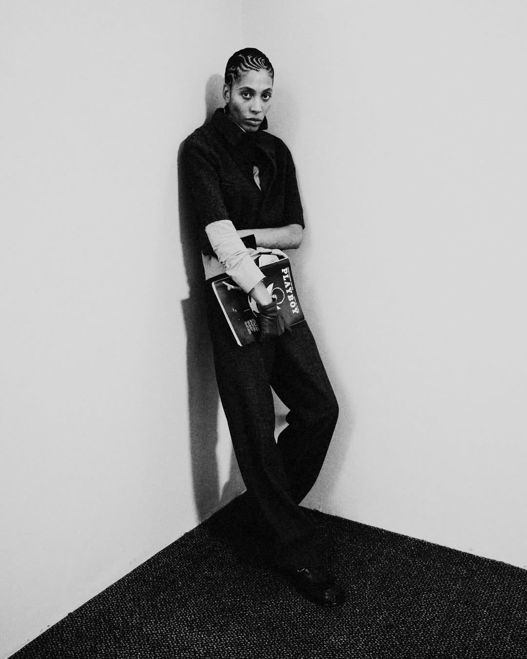 Black and white portrait of a woman with braided hair leaning against a corner holding a magazine, showcasing fashion and style.