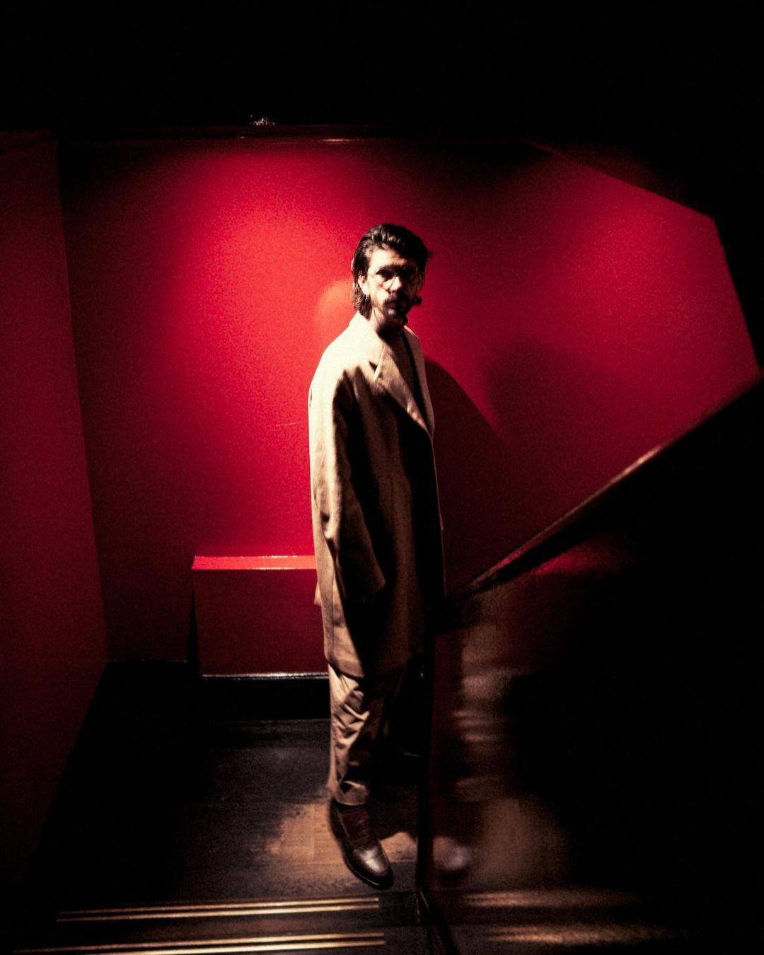 Man in suit jacket walking down stairs, bathed in moody red and gold light, creating an ambiance of drama and intrigue.