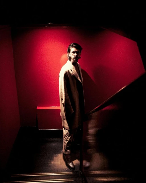 Man in suit jacket walking down stairs, bathed in moody red and gold light, creating an ambiance of drama and intrigue.