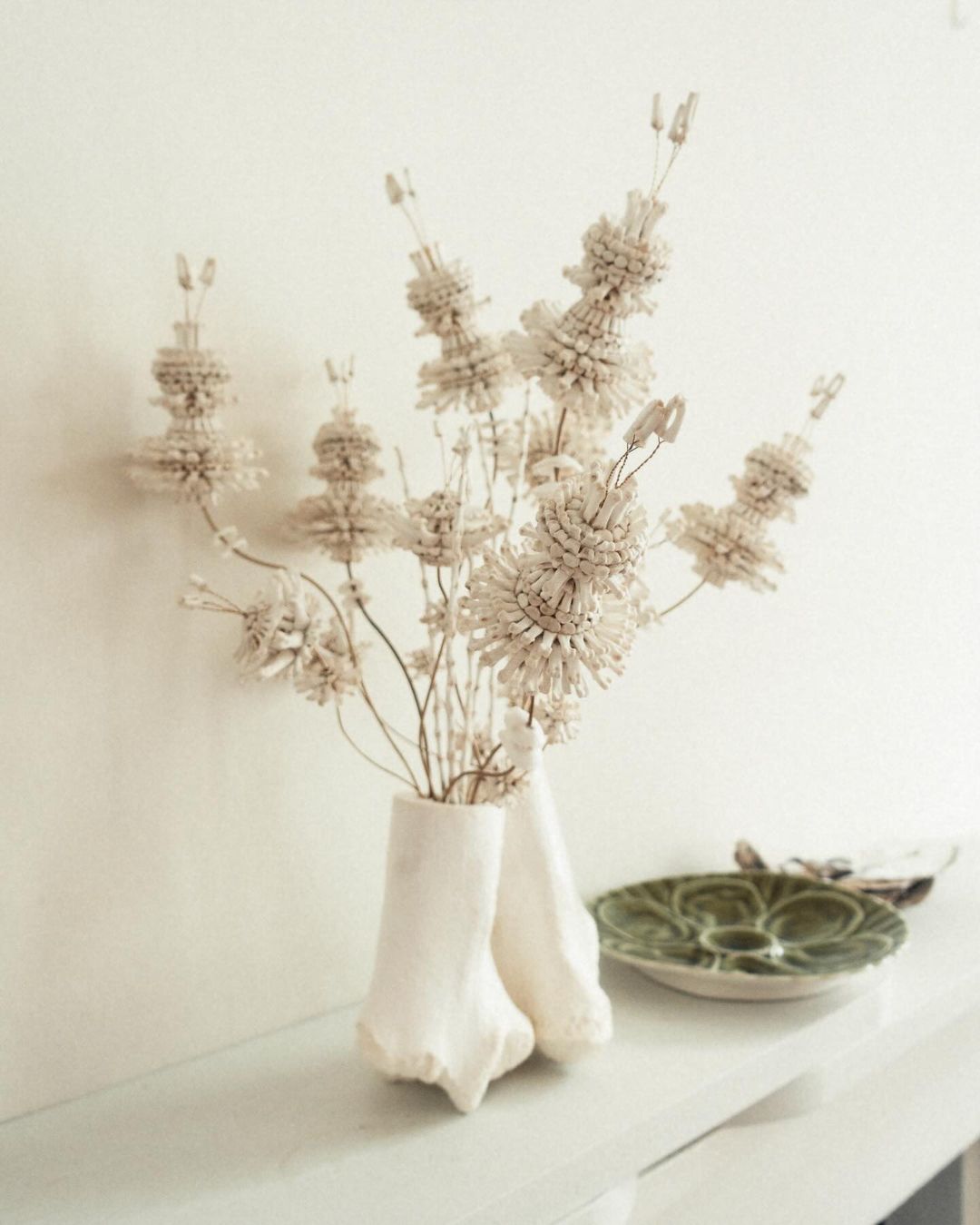 A vase with dry, decorative floral arrangements against an off-white wall, with a green ceramic plate on a shelf in the background.