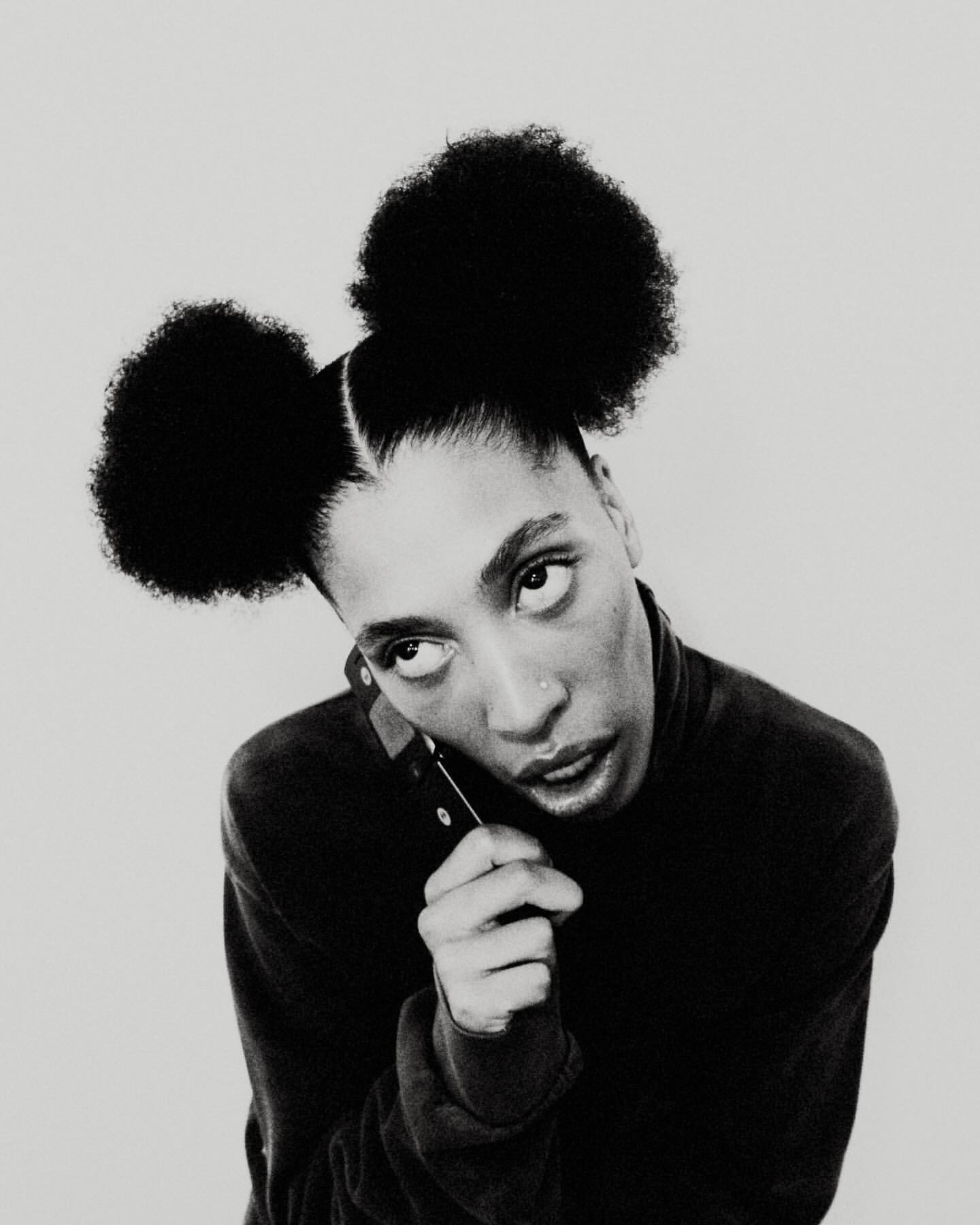 Portrait of an adult female with afro puffs using a cell phone against a neutral background. The model is wearing a black turtleneck sweater.