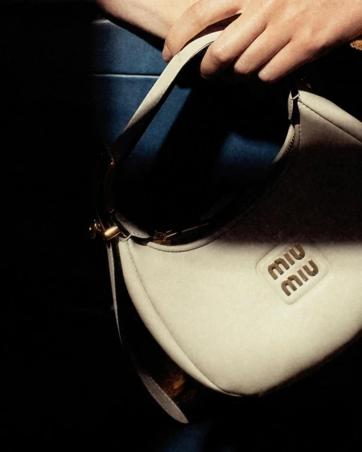 Close-up of a hand holding a Miu Miu ivory-colored leather handbag against a dark blue and black backdrop in a studio setting.