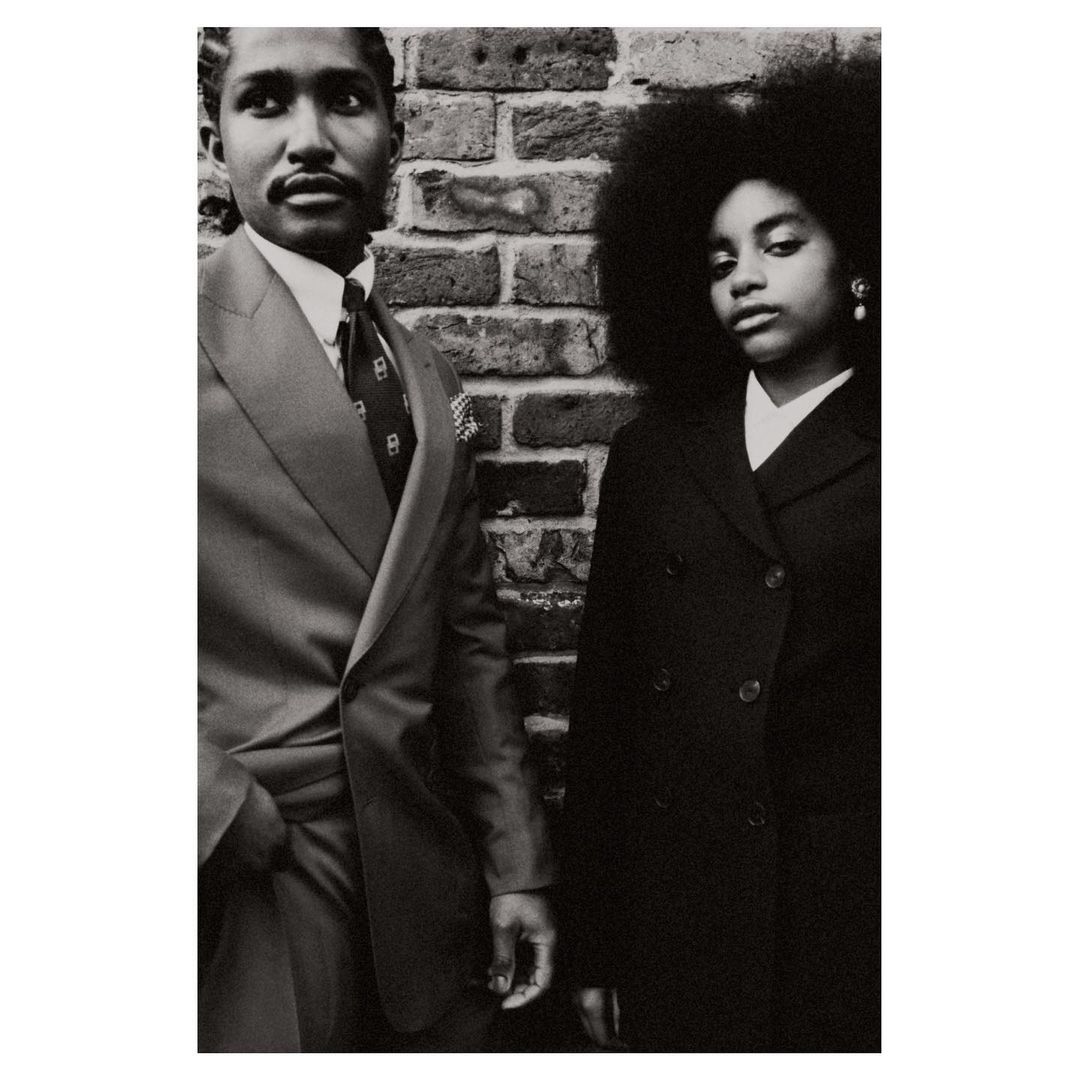 Black and white portrait of an African American couple in suits standing against a brick wall, exuding vintage fashion and style.