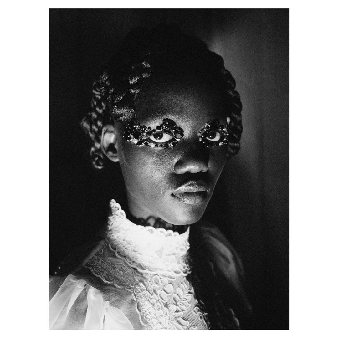 Monochrome portrait of a young African woman wearing elaborate eye makeup and a lacy white dress with braided hair in a dark, dramatic setting.