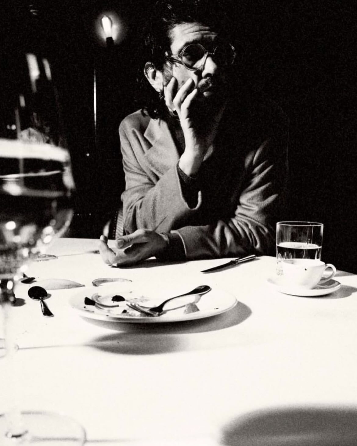 Monochromatic photograph of a man with glasses sitting at a table with dishes, silverware, a glass of water, and a white tablecloth.