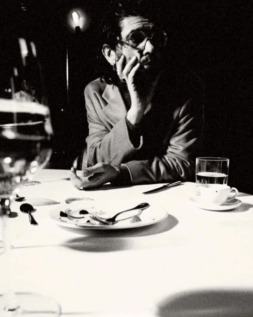 Monochromatic photograph of a man with glasses sitting at a table with dishes, silverware, a glass of water, and a white tablecloth.