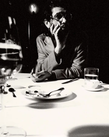 Monochromatic photograph of a man with glasses sitting at a table with dishes, silverware, a glass of water, and a white tablecloth.