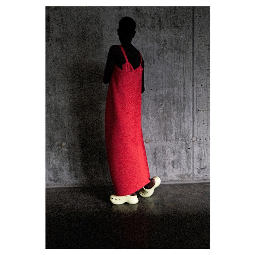 Model in a long red dress and off-white clog shoes stands against a textured grey wall in a studio setting with dark lighting.