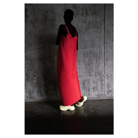 Model in a long red dress and off-white clog shoes stands against a textured grey wall in a studio setting with dark lighting.