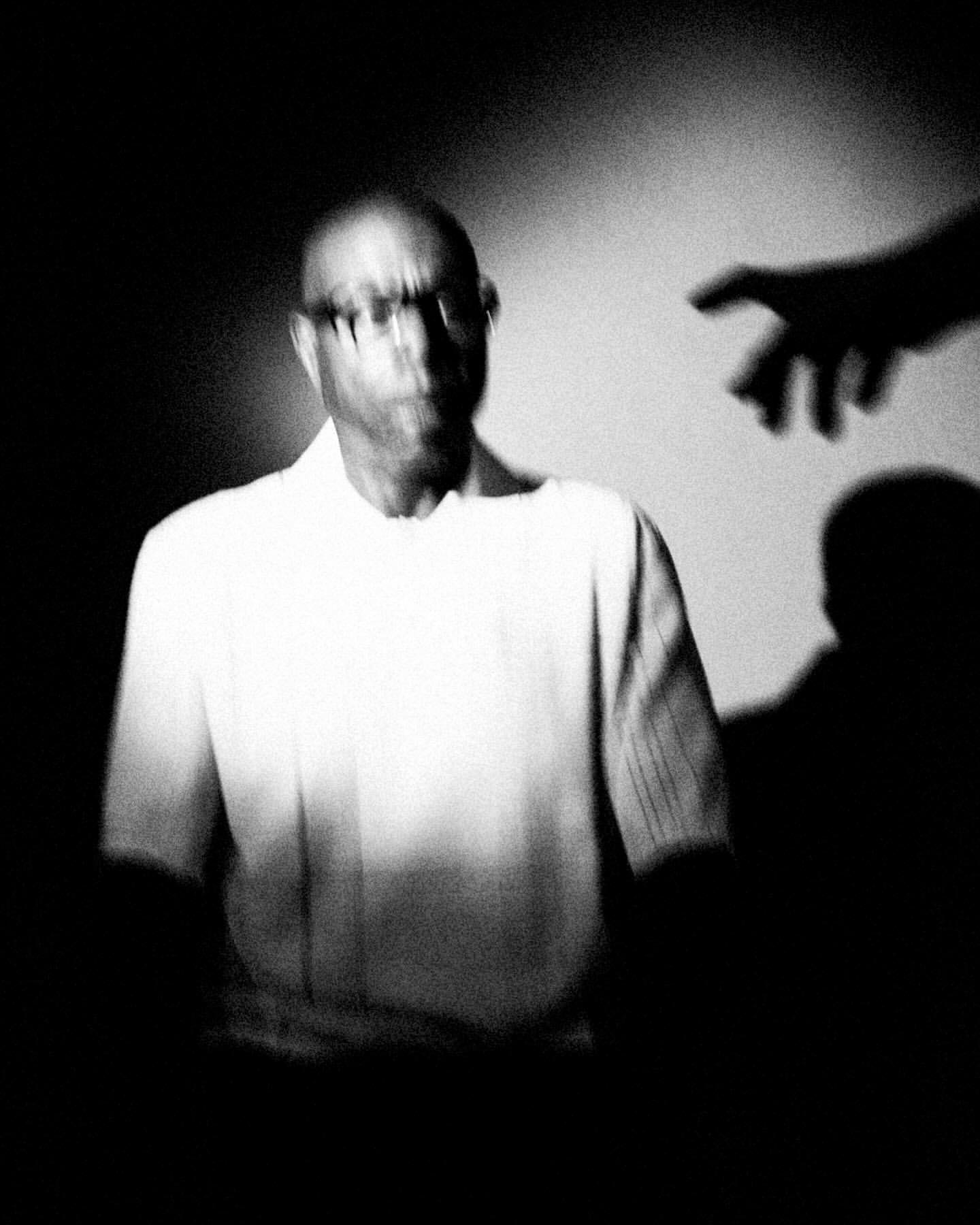 Monochrome portrait of a bald, bespectacled man with a pointing hand shadow, creating a stark, high-contrast image against a plain backdrop.