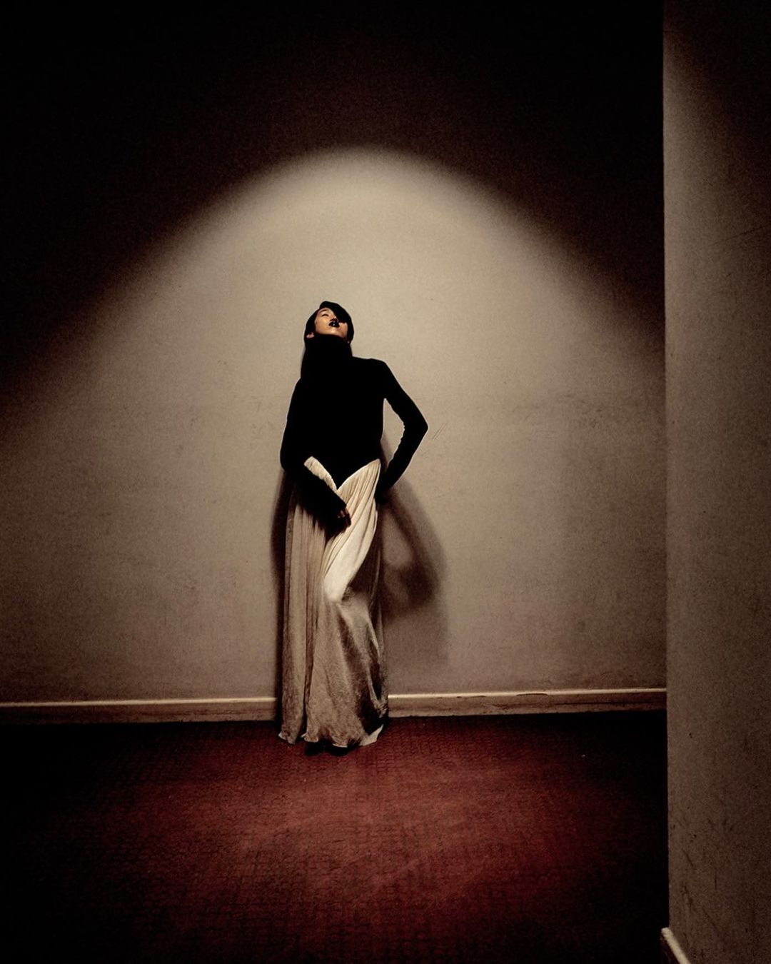 An adult female fashion model in a black turtleneck and long flowing skirt is posing dramatically in a dimly lit interior space.