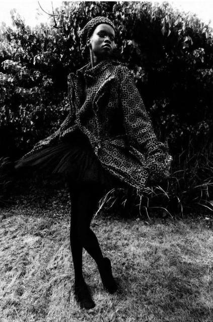 Monochrome full shot of a slender woman in a printed coat and beret, posing outdoors with a blurred, leafy background.