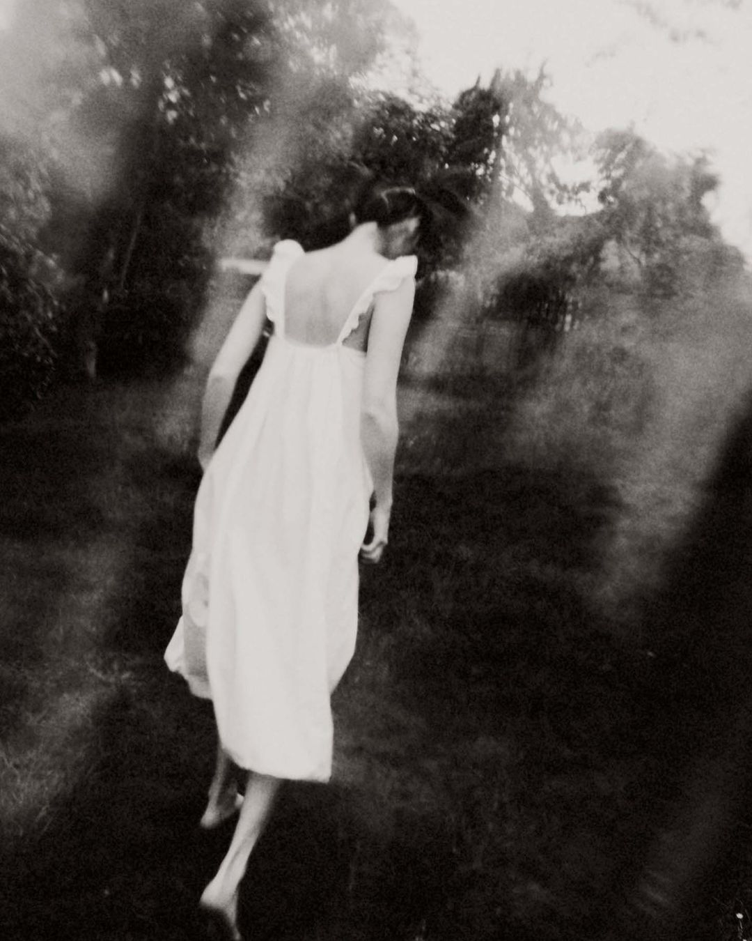 Woman in a long white dress walking through a dark garden captured in blurry monochromatic style